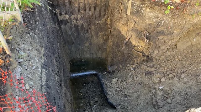 Excavation site with deep trench dug in dark earth, showing exposed soil layers, water and corrugated plastic drainage pipe