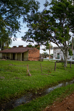 View of a weathered brick building with a rusted roof set in a grassy field with a drainage ditch and large trees in Guaviravi, Corrientes Province, Argentina.