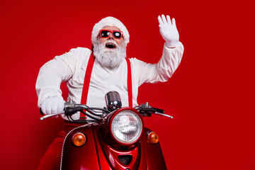 Obraz premium Santa Claus riding a red scooter wearing sunglasses and waving — festive Christmas portrait with white beard, gloves and suspenders on a bold red background