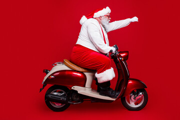 Obraz premium Santa Claus riding a red vintage scooter in festive costume on a red background, playful holiday transportation and Christmas celebration scene