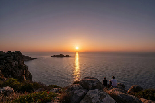 Two people sitting on rocky coastal cliffs looking out at a sunset over the ocean with sunset landscape