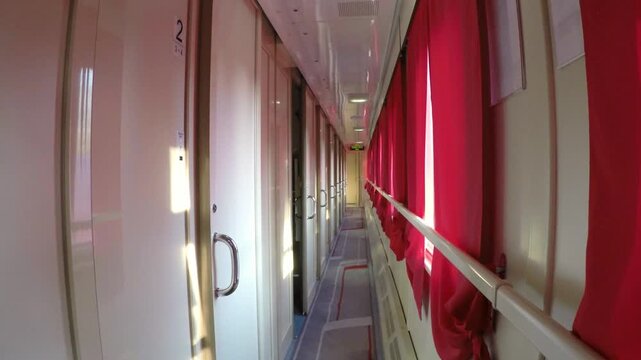 Interior view along the narrow corridor of a moving passenger train carriage with red curtains on the windows, compartment doors, and dynamic sunlight flashing through the glass during travel.