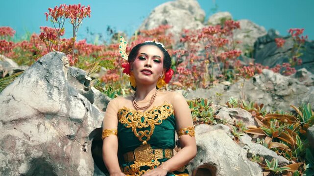 A portrait of a traditional Javanese bride in a green costume, sitting with poise in a stone garden, looking towards the camera.