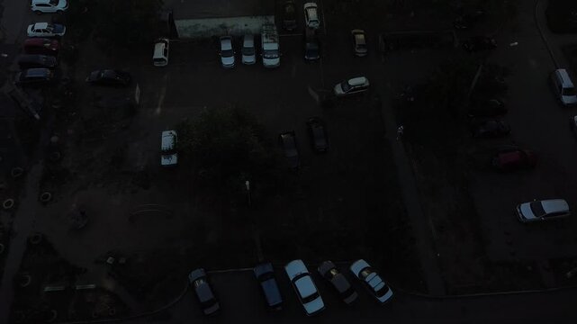 Aerial view captures urban streets at dusk. Shadows stretch across rooftops and pavement. Vehicles idle near construction sites and alleys. People move quietly, unseen from above