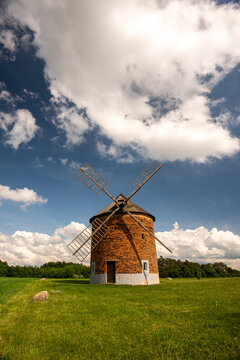 View of a historic brick windmill standing in a green field under a bright blue sky with fluffy white clouds in Brno-City District, South Moravian Region, Czechia.