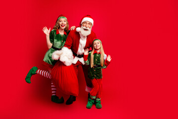 Obraz premium Santa Claus with two cheerful elves posing with a large red gift sack in festive green and red costumes — family holiday portrait conveying joyful Christmas celebration