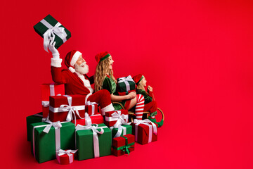 Obraz premium Santa Claus and two festive elves sitting among wrapped Christmas presents on a red background, celebrating holiday joy, family fun and playful seasonal cheer