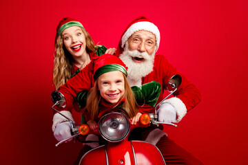 Obraz premium Santa and two cheerful elves riding a red scooter for Christmas, smiling family holiday portrait with festive hats and joyful expressions