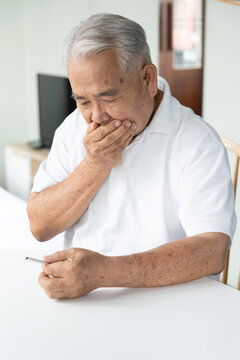 70s senior southeast asian man holding a cigarette at home, feeling sick and regretful about smoking indoors, concept image for quit smoking awareness