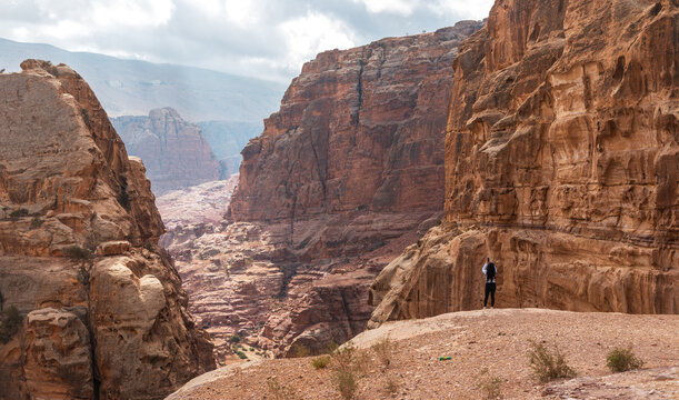 View of a hiker standing on a rocky ledge overlooking the deep, rugged sandstone canyon of Wadi Musa, Ma'an Governorate, Jordan.
