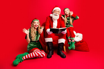 Obraz premium Santa Claus reading with two cheerful elves and children on a red background for Christmas holiday family portrait and festive storytelling