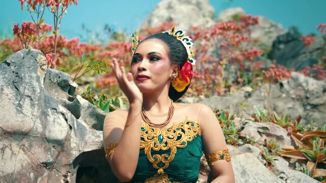 Footage of an Indonesian dancer in a green outfit performing graceful traditional hand movements (mudras) while sitting against a rocky backdrop.