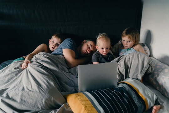 Three little children are watching a laptop while their exhausted father is sleeping beside them in a cozy bed. This authentic family lifestyle concept highlights modern parenting struggles.
