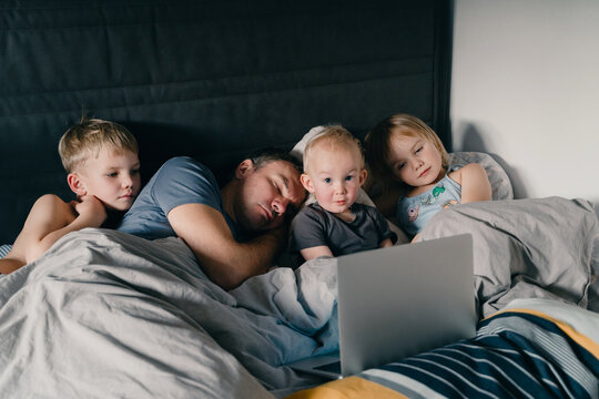 Three little children are watching a laptop while their exhausted father is sleeping beside them in a cozy bed. This authentic family lifestyle concept highlights modern parenting struggles.