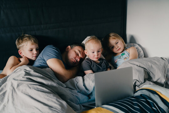 Three little children are watching a laptop while their exhausted father is sleeping beside them in a cozy bed. This authentic family lifestyle concept highlights modern parenting struggles.