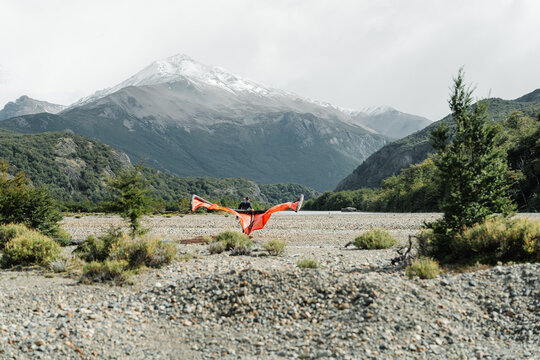 View of a person holding an orange wing in a rocky riverbed with snow-capped mountains in the background in El Chalten, Santa Cruz Province, Argentina.