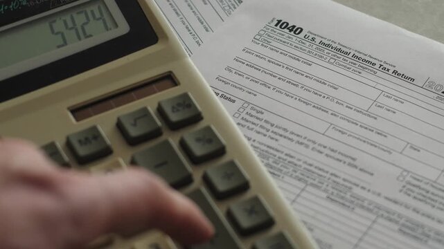 A person presses buttons on a desktop calculator while calculating numbers near a US Form 1040 individual income tax return. An accountant conducts a financial audit and plans the budget