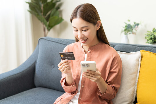 Bill pay, shopping order online internet, cute asian young woman hand holding credit card and mobile smart phone on sofa at home, female using banking service to secure payment with purchase on store.