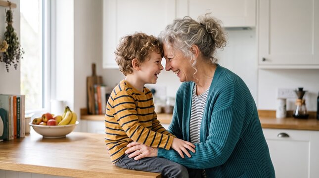 Smiling senior woman and small curly‑haired boy touch foreheads at a kitchen island, hands on each other&rsquo;s arms, with fruit bowl and cookbooks on the counter