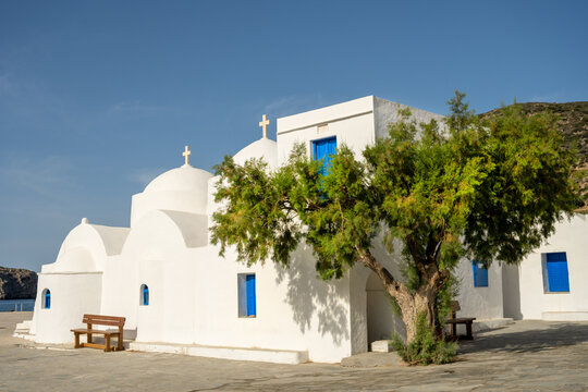 View of a traditional white-washed Greek Orthodox church with blue doors and domes, featuring a large tree in the foreground under a clear blue sky in Lefkada, Greece.
