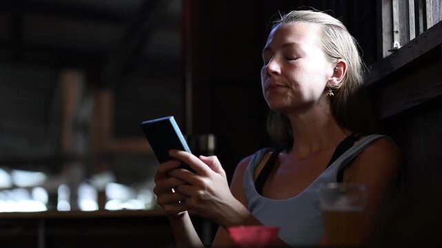 Relaxed caucasian woman using her mobile phone, scrolling social media and texting while sitting in a traditional wooden bar in Chiang Mai, Thailand, during her vacation