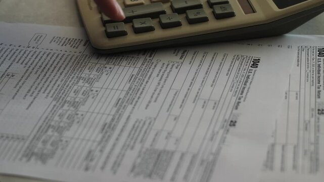 US individual income tax return form 1040 and a calculator are placed on a desk for financial accounting. A person presses buttons on the calculator while auditing documents 