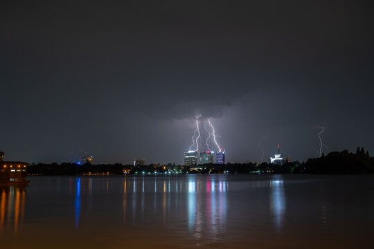 View of lightning strikes illuminating the night skyline across the lake with reflections on the water surface in Bucharest, Bucharest, Romania.