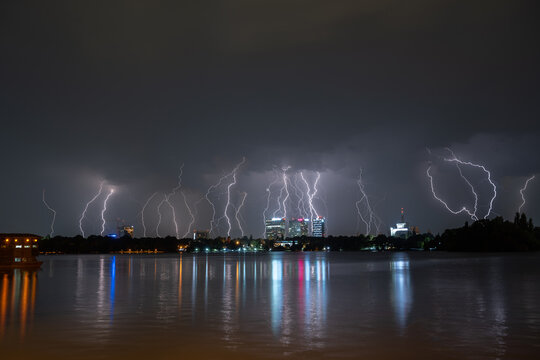 View of multiple lightning bolts striking the night sky over the illuminated Bucharest skyline reflected in the calm waters of Herastrau Lake, Bucharest, Bucharest, Romania.