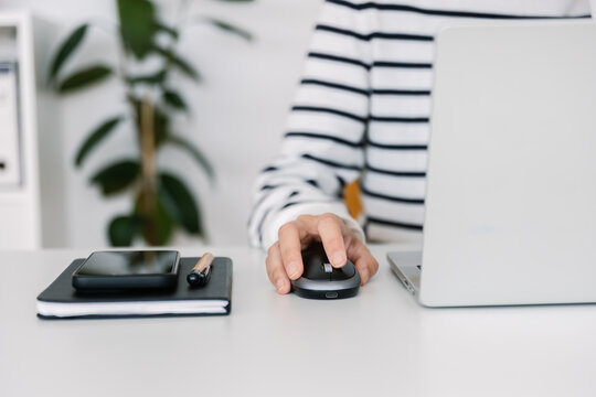 Close up of woman hand using computer mouse next to laptop at home office. Unrecognizable female freelancer working at desk with notebook and phone. Technology and business concept.