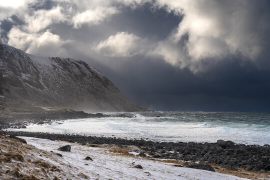 View of a snowy coastal landscape with a rocky beach, crashing waves, and dramatic dark storm clouds over the mountains in Leknes, Lofoten, Norway.