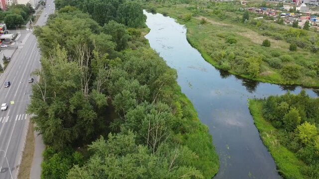 Aerial view captures a winding river bordered by lush greenery. Riverbanks are lined with tall trees and thick undergrowth. Road runs parallel to the water, with cars moving steadily along