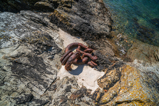 View of a heavy rusty metal chain resting on jagged coastal rocks beside the clear water of the Fowey River in Fowey, Cornwall, United Kingdom.