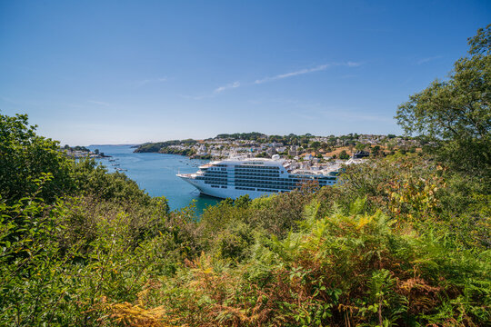 View of a large cruise ship anchored in the Fowey River with the town of Fowey in the background, framed by lush trees and ferns under a clear blue sky in Fowey, Cornwall, United Kingdom.