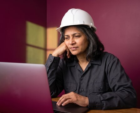 Pensive civil engineer in white hardhat resting chin on hand beside laptop