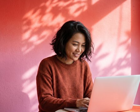 Curious creative woman in rust sweater working on laptop against pink wall with leaf shadow