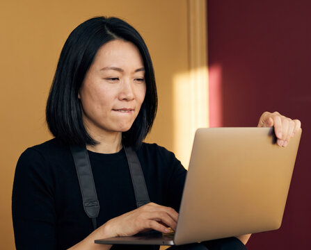 Asian female photographer in black apron biting lip while holding laptop against mustard wall
