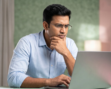 Analytical man in glasses and blue shirt frowning in concentration while working on laptop
