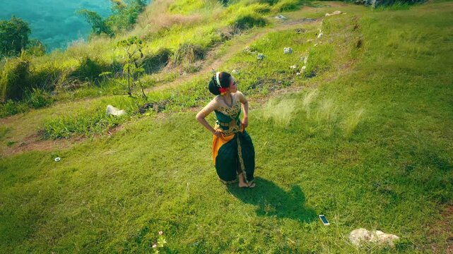 High angle footage of a female dancer in traditional attire executing dynamic dance moves on a grassy terrain surrounded by rocks.