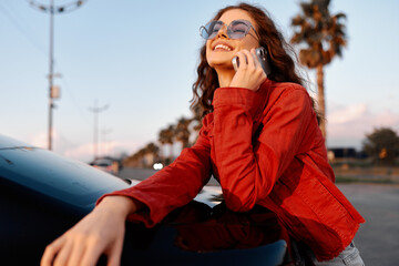 Naklejka na ściany i meble Young woman in a red jacket smiling while talking on her phone outdoors. She leans against a car during sunset. Lifestyle concept for modern communication, travel, and urban lifestyle.