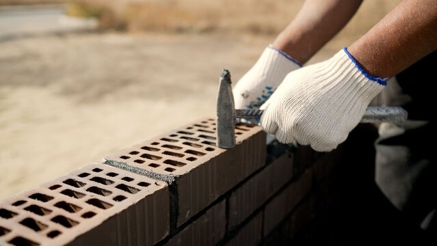 Worker in white gloves using hammer to lay brown perforated bricks on wall at construction site, masonry and building trade