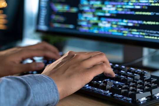 Hands type code on a glowing keyboard. Blue backlight illuminates fingers and keys. Monitor displays lines of programming syntax. Focus suggests deep concentration or problem-solving