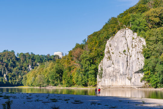 Two hikers on the gravel bank of the Danube River beneath a white limestone cliff in the Danube Gorge near Kelheim, Lower Bavaria, Germany, Liberation Hall on the hilltop, sunny autumn day