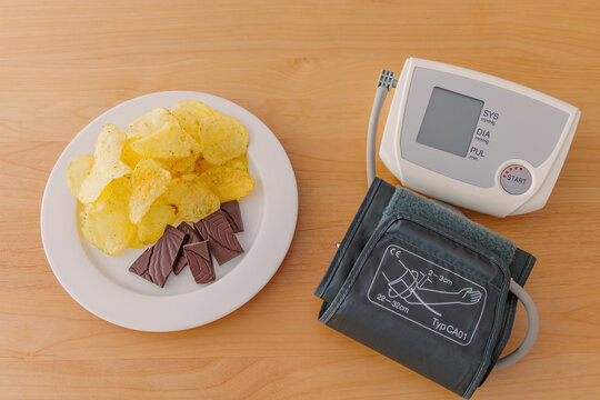 Plate of potato chips and dark chocolate next to a blood pressure monitor with cuff on wooden table, unhealthy diet, hypertension and cardiovascular risk concept, top view