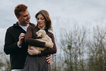 Obraz premium Couple holds a goat kid in their arms outdoors, sharing affection and a joyful moment, showcasing rural lifestyle and tender bond in a natural, warm scene.