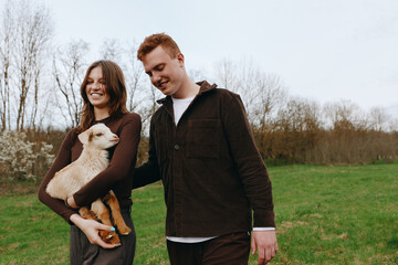 Obraz premium Joyful couple outdoors with a goat kid, walking through a green field, warm light, smiling faces, relaxed posture, natural tones, carefree moment, countryside vibe, gentle breeze vibrant