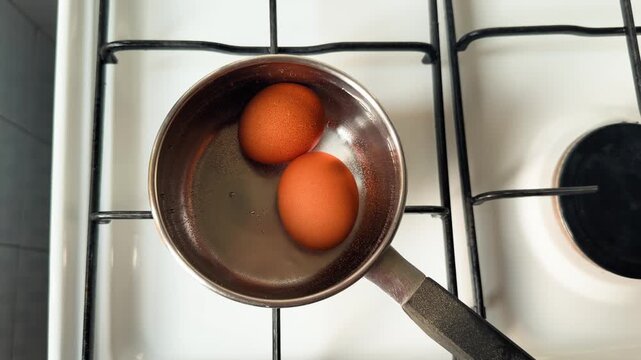 Close up of two brown eggs in a small metal saucepan on a kitchen stove, viewed from above.