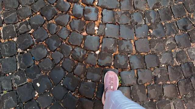 Close up of a person walking on old uneven cobblestone pavement, showing worn stone texture and a casual urban scene.