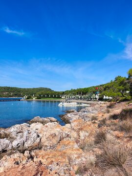 Scenic view of Ortunc Cove in Cunda Island, Ayvalik with crystal clear sea, pebble beach, pine trees, and a small boat under a blue sky, Balikesir, Turkey.