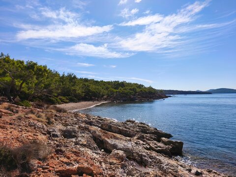 Scenic view of Ortunc Cove in Cunda Island, Ayvalik with crystal clear sea, pebble beach, pine trees, and a small boat under a blue sky, Balikesir, Turkey.