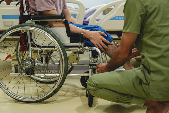 Caregiver or male physiotherapist assisting disabled man sitting in wheelchair, checking leg during physical therapy in hospital room.